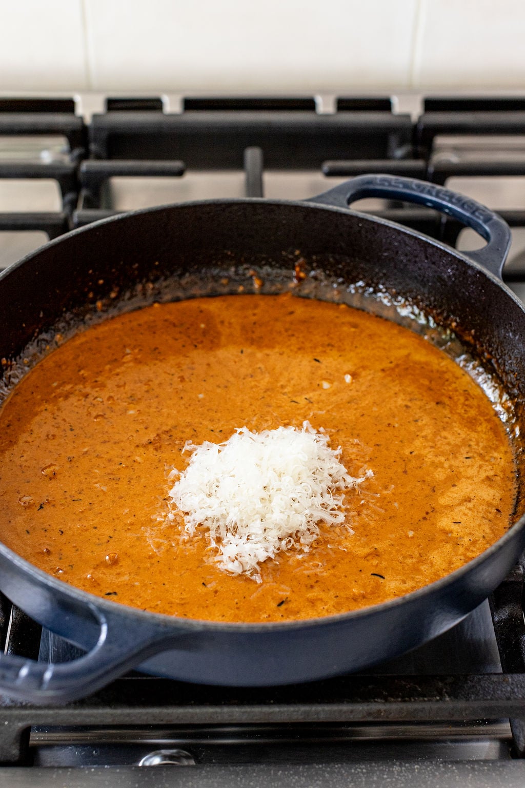 A black skillet on a stovetop contains a reddish-brown sauce with a small mound of grated cheese in the center.