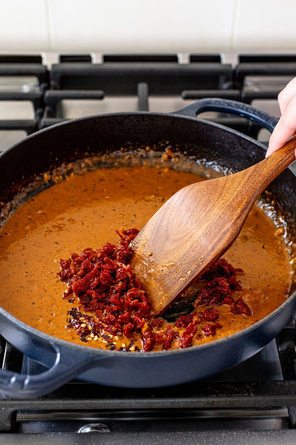 A wooden spatula stirs chopped sun-dried tomatoes into a creamy brown sauce in a black skillet on a stovetop.