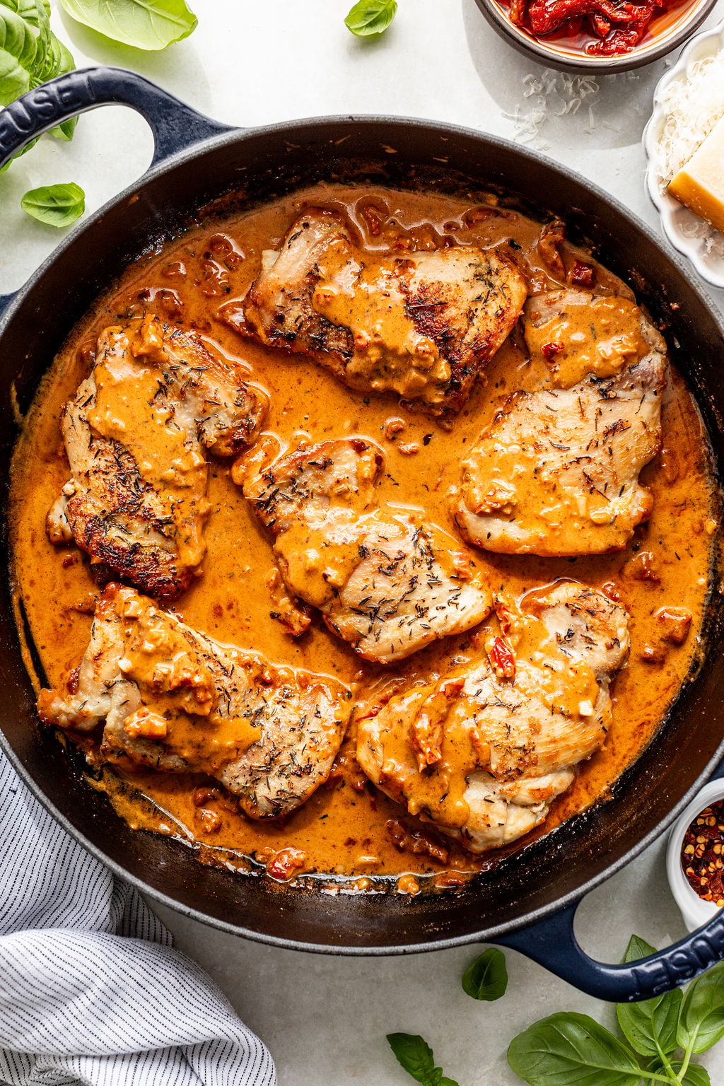 Chicken breasts cooked in a creamy, orange-colored sauce with herbs and sun-dried tomatoes in a black skillet. Fresh basil leaves and ingredients are placed around the pan.