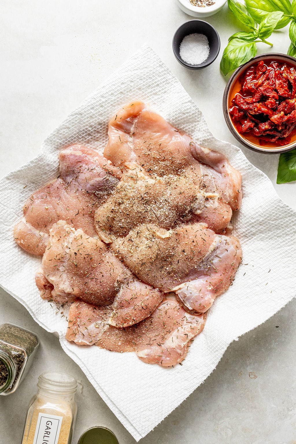 Raw, seasoned chicken breasts placed on paper towels, surrounded by bowls of sun-dried tomatoes, basil leaves, salt, and various seasoning containers on a light surface.