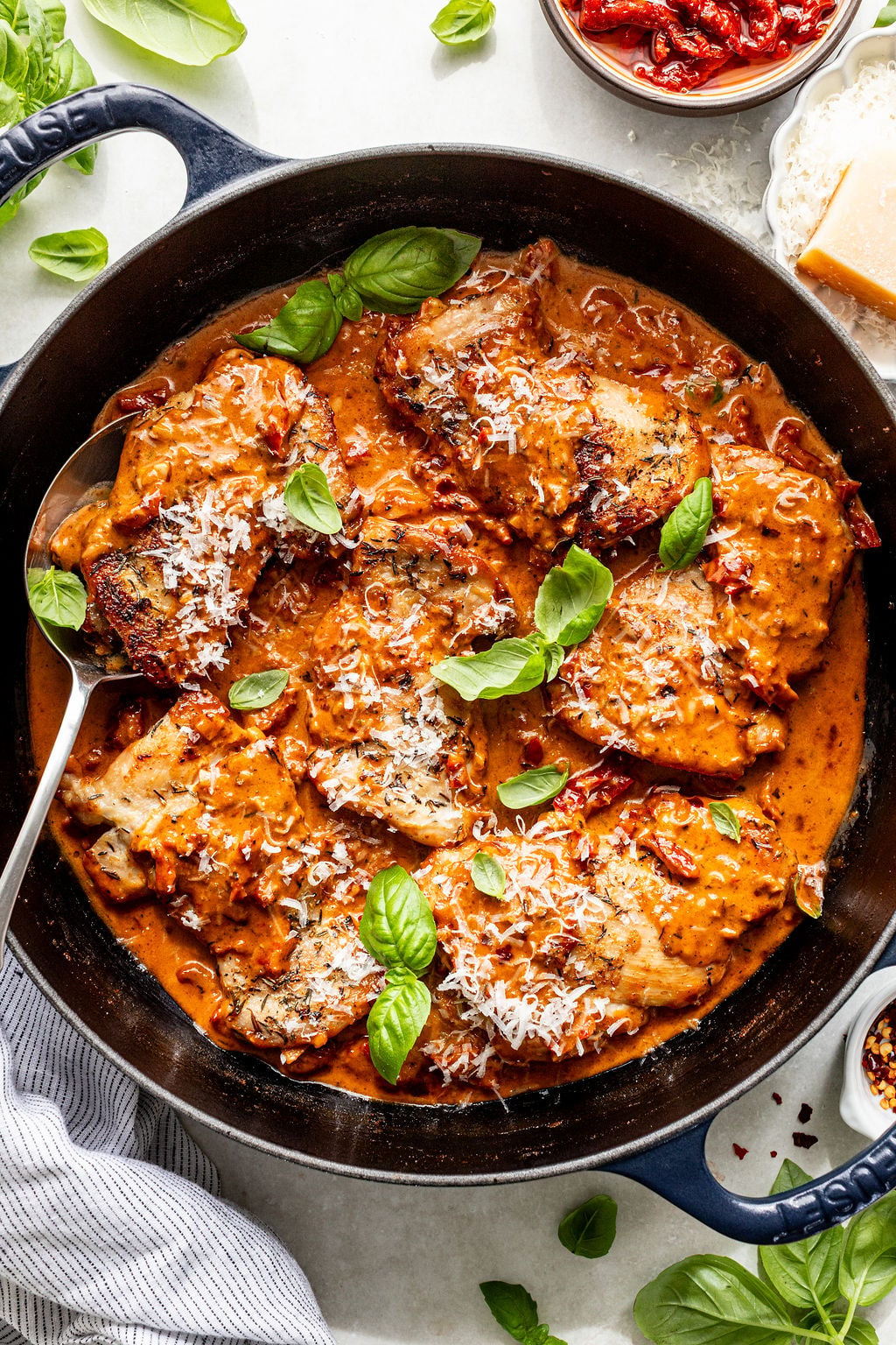 Skillet with chicken in a creamy tomato sauce, topped with grated cheese and fresh basil leaves, with a serving spoon.