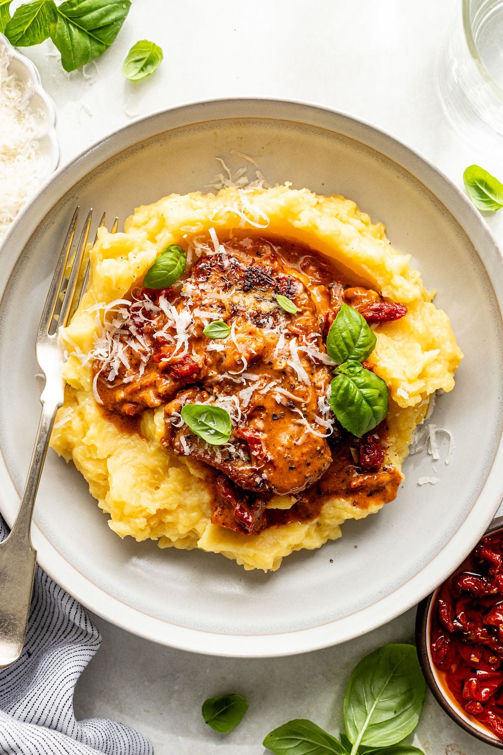 A plate of mashed potatoes topped with saucy chicken, sun-dried tomatoes, grated cheese, and fresh basil leaves, with a fork on the side.