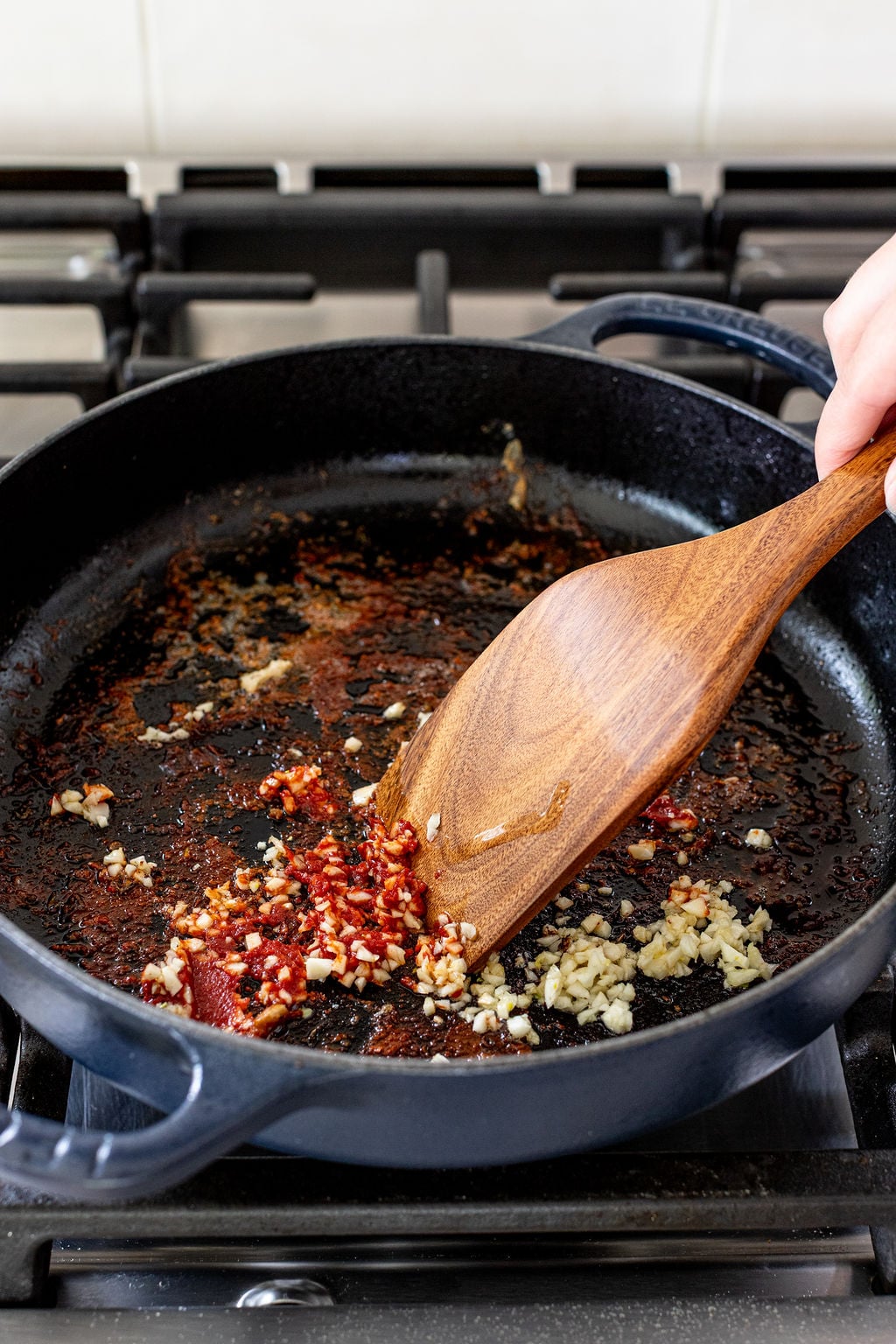 A hand stirs minced garlic and chili flakes in a black skillet with a wooden spatula on a stovetop.