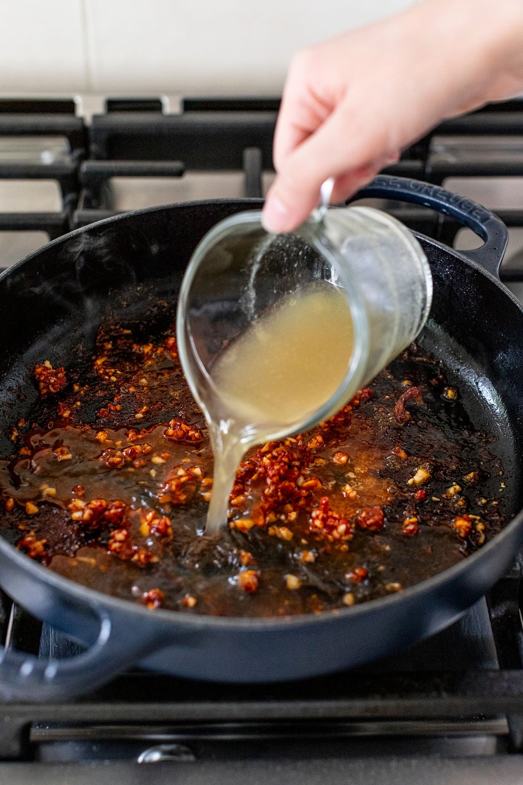 A hand pours broth from a glass measuring cup into a skillet containing sautéed garlic and chili flakes on a stovetop.