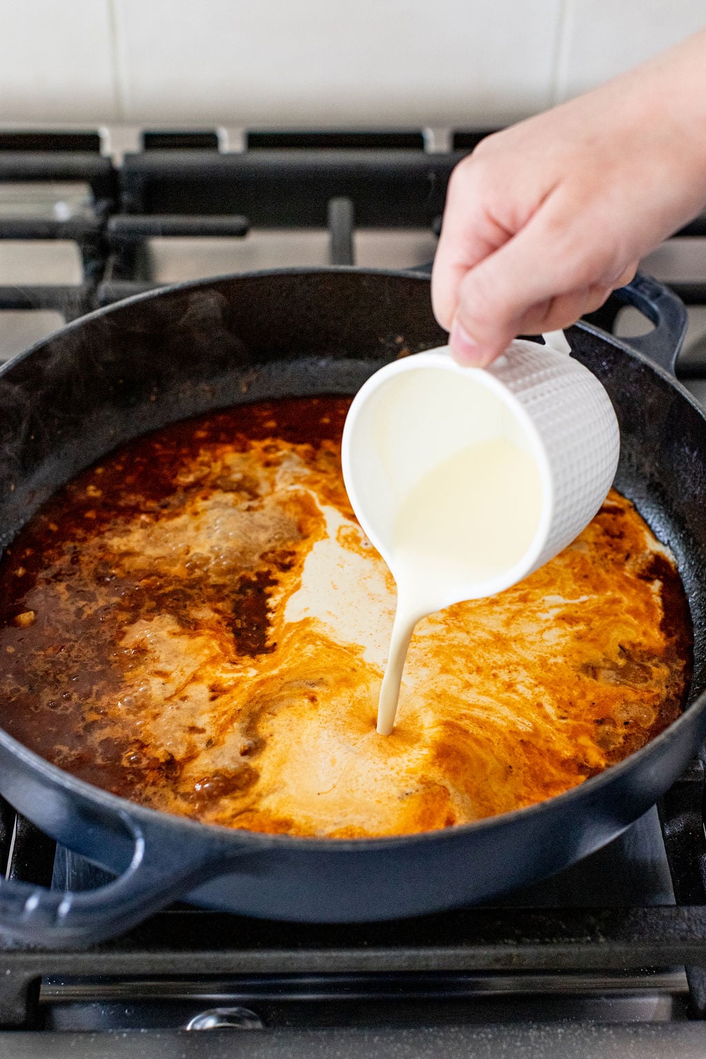 A hand pours cream from a small white pitcher into a black skillet with a dark, simmering sauce on a stove.
