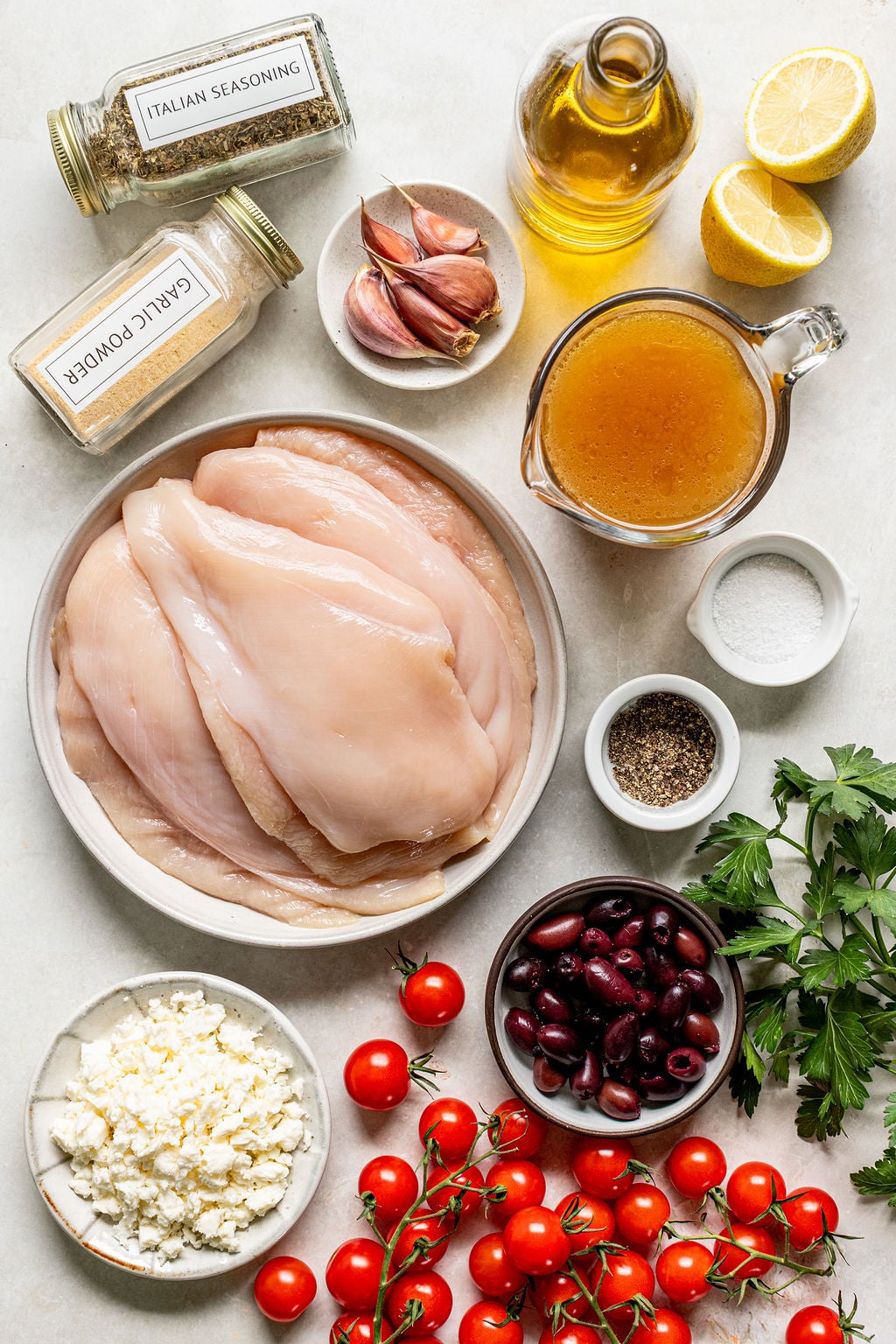 Raw chicken breasts surrounded by Italian seasoning, garlic, olive oil, lemon halves, broth, salt, pepper, parsley, Kalamata olives, feta cheese, and cherry tomatoes on a white surface.