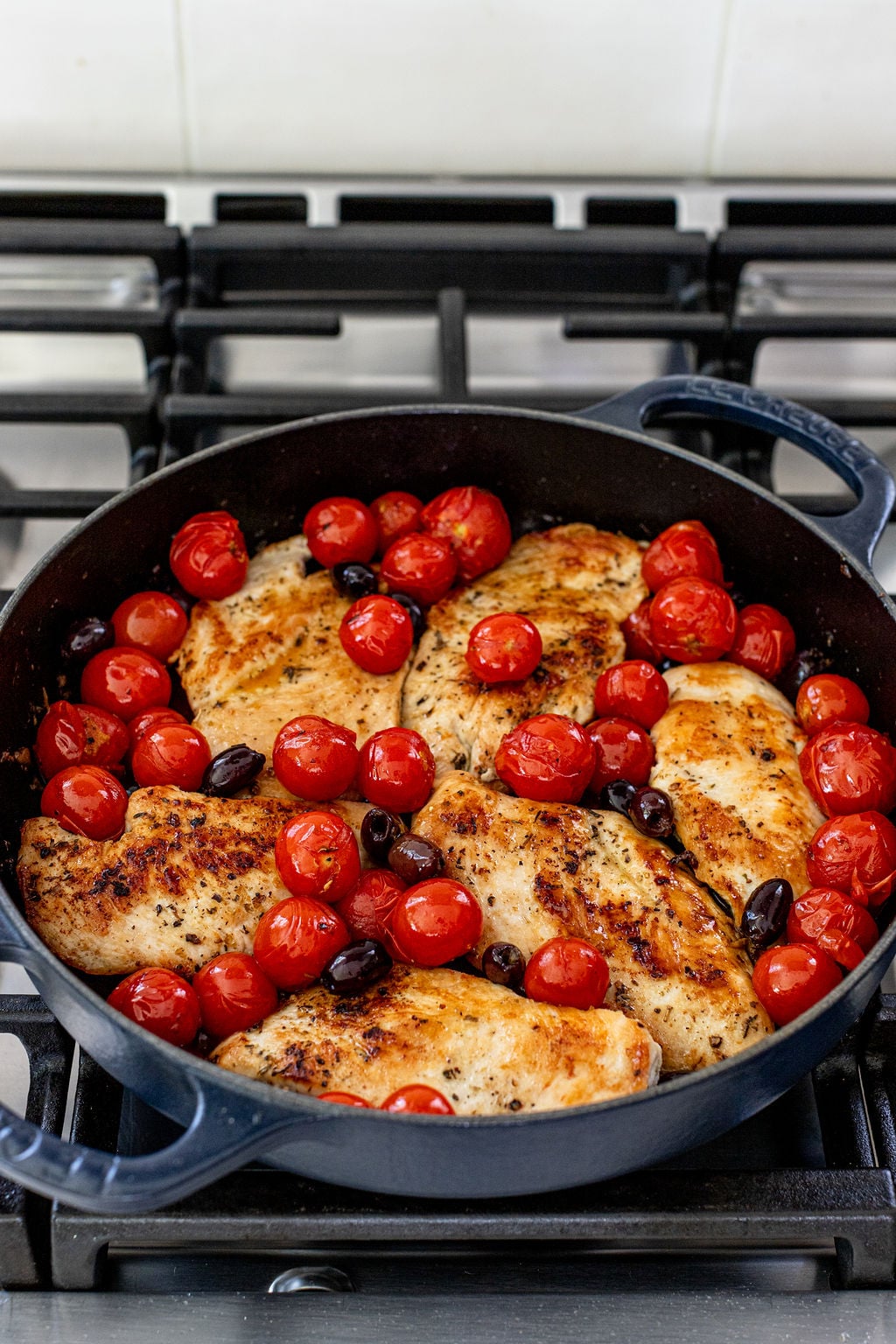 Chicken breasts, cherry tomatoes, and black olives cooking in a black skillet on a stovetop.