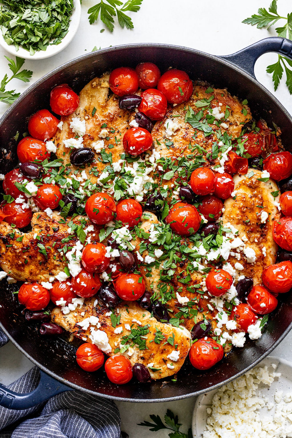 Skillet with cooked chicken breasts, roasted cherry tomatoes, kalamata olives, crumbled feta cheese, and chopped parsley, with a bowl of chopped herbs on the side.