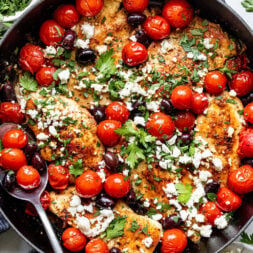 A skillet filled with cooked chicken breasts topped with cherry tomatoes, olives, crumbled feta cheese, and fresh parsley. A serving spoon rests inside the skillet.