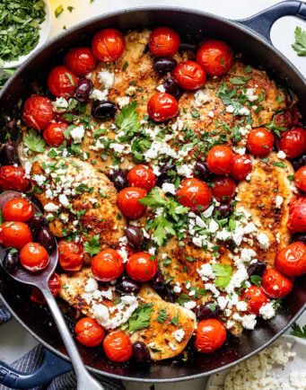 A skillet filled with cooked chicken breasts topped with cherry tomatoes, olives, crumbled feta cheese, and fresh parsley. A serving spoon rests inside the skillet.