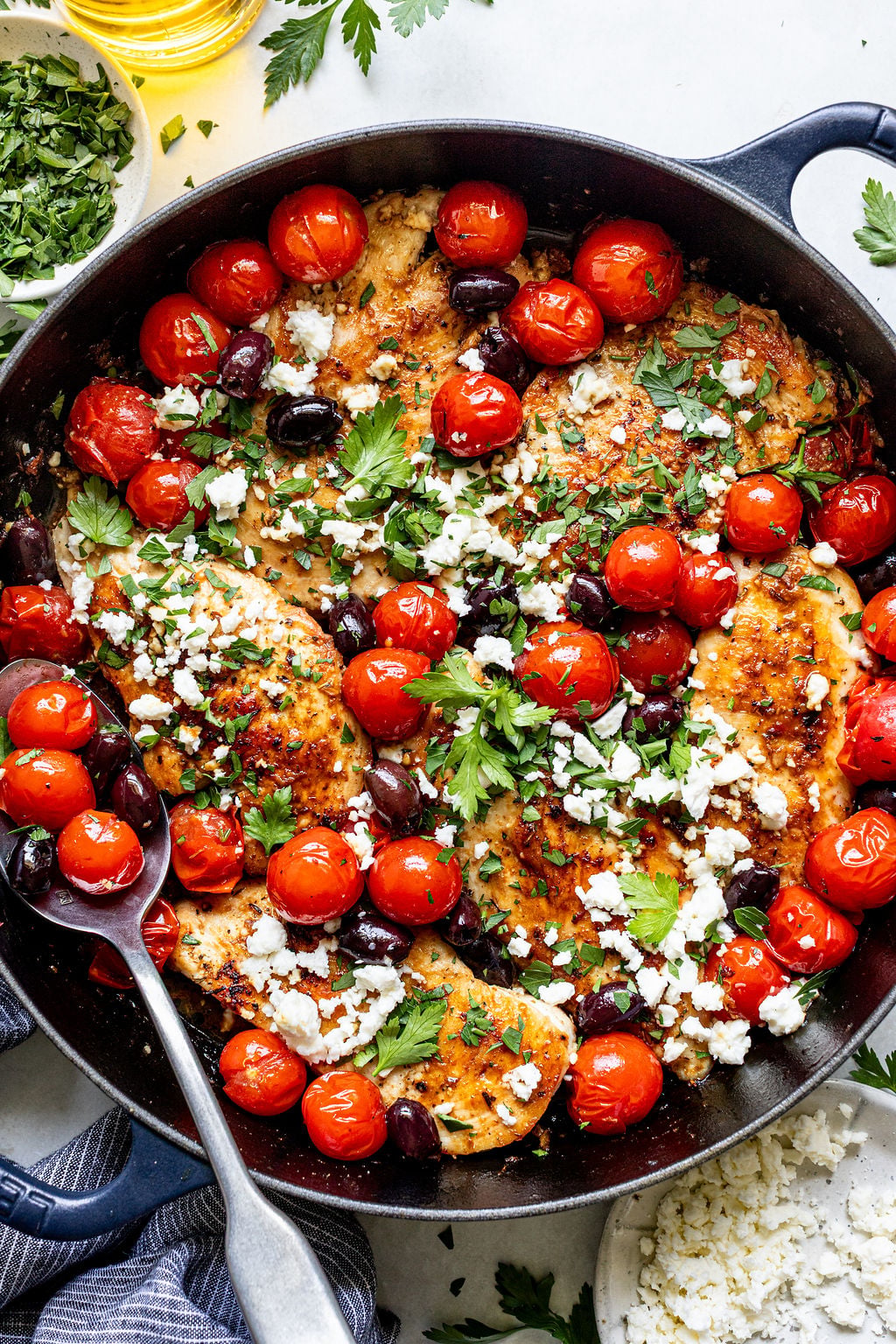 A skillet filled with cooked chicken breasts topped with cherry tomatoes, olives, crumbled feta cheese, and fresh parsley. A serving spoon rests inside the skillet.