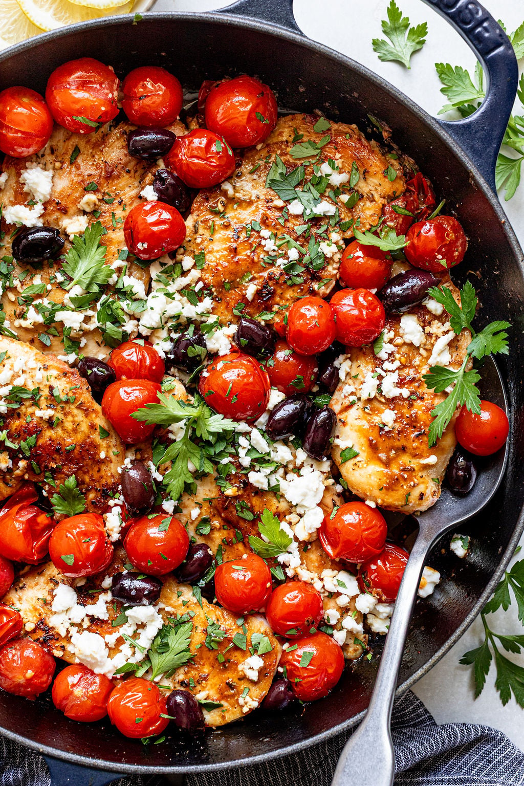 A skillet with cooked chicken breasts topped with cherry tomatoes, black olives, crumbled feta cheese, and fresh parsley.