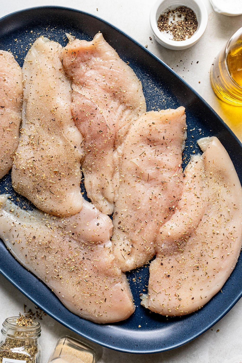Raw, seasoned chicken breast fillets are arranged on a dark oval plate. Various spices and a small bowl of pepper are visible nearby.