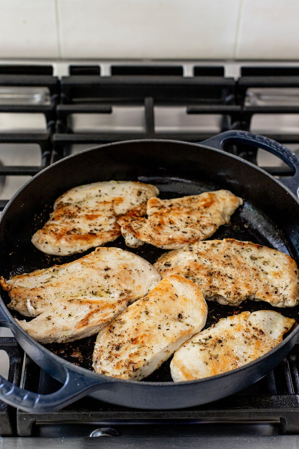 Six seasoned chicken breasts cooking in a black skillet on a gas stove.