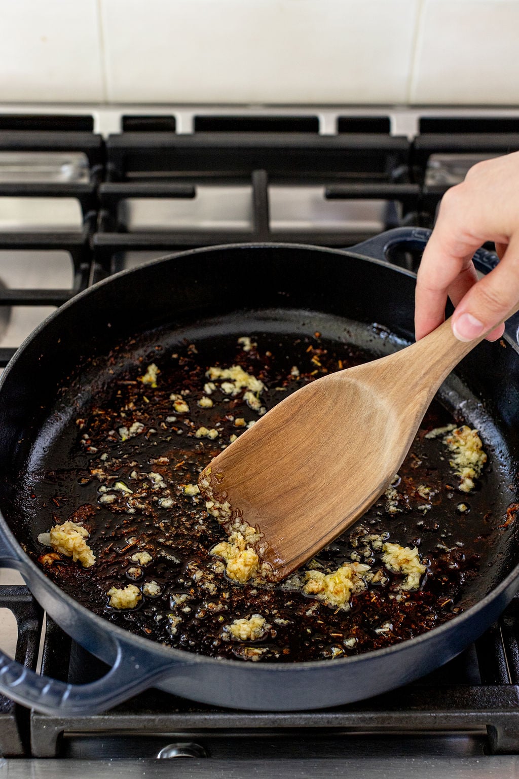 A hand uses a wooden spatula to saut&eacute; minced garlic in a black skillet on a stovetop.