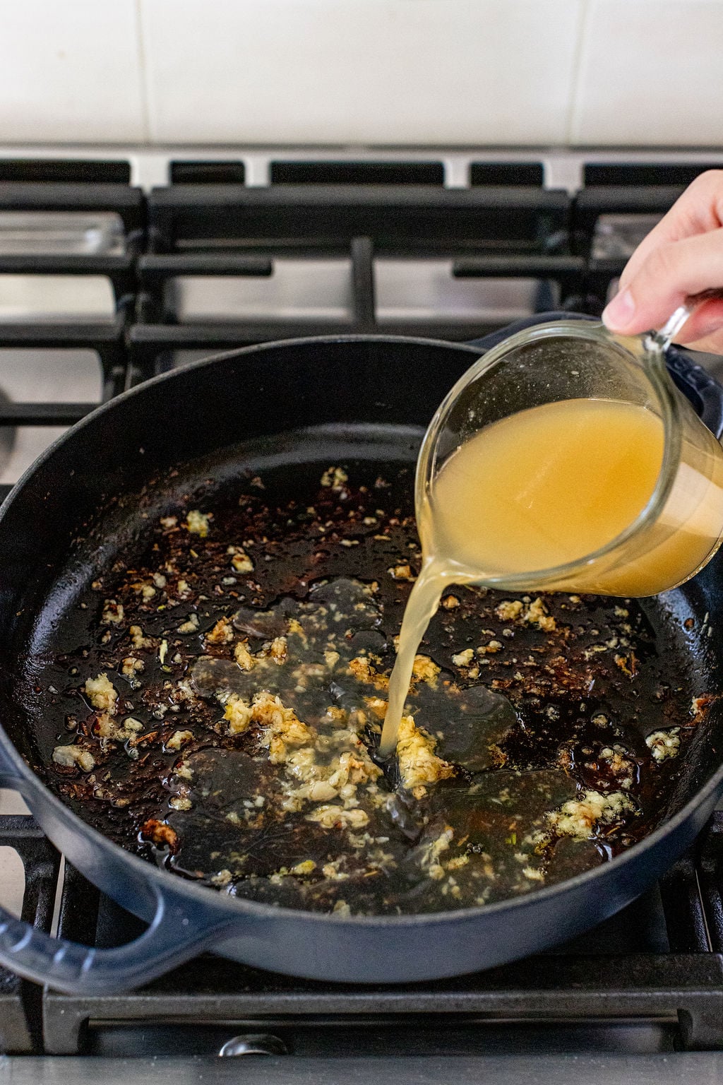 A hand pours broth from a measuring cup into a black skillet with saut&eacute;ed garlic and browned bits on a stovetop.