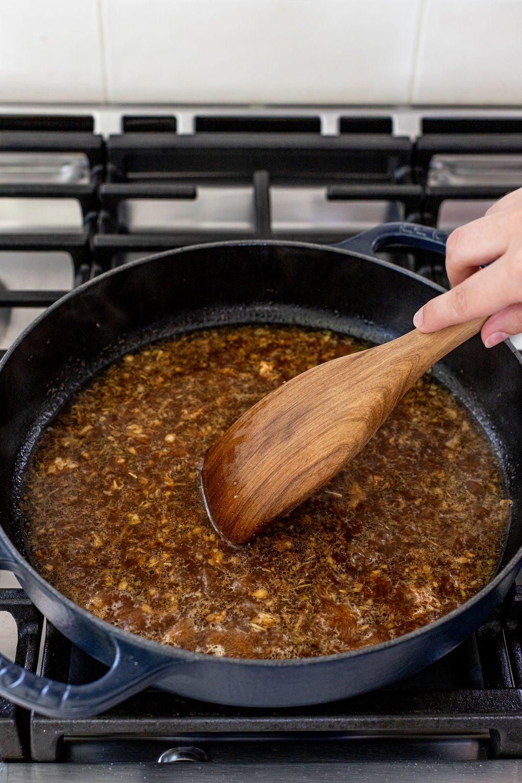 A hand stirs a brown liquid in a black pan with a wooden spoon on a gas stove.