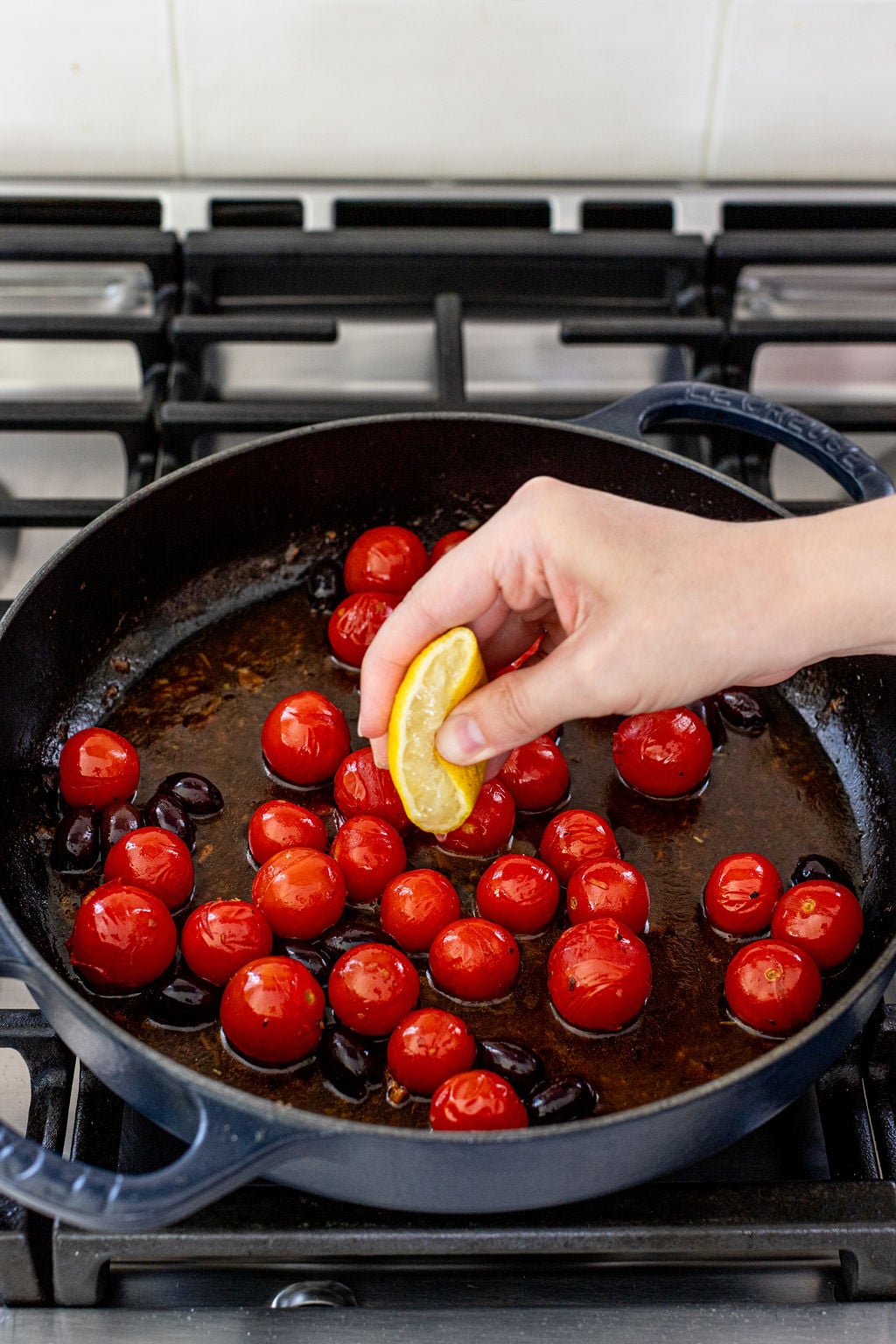 A hand squeezing a lemon wedge over a skillet of cherry tomatoes and black olives cooking on a stovetop.