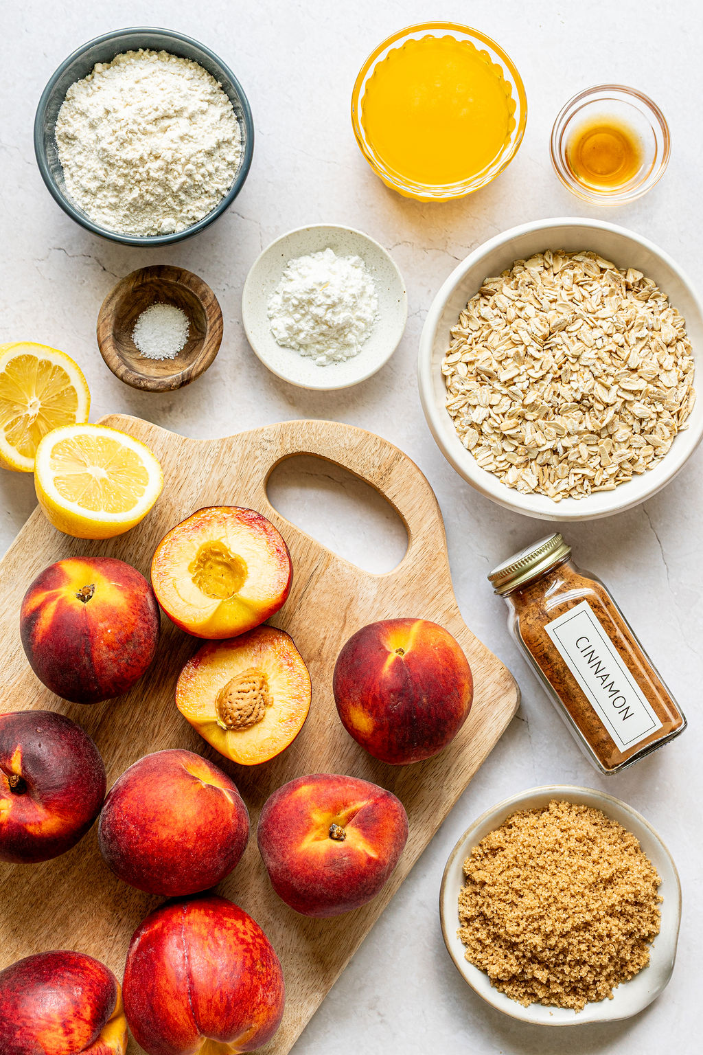 Various ingredients for baking, including fresh peaches, oats, brown sugar, flour, salt, cinnamon, lemon, orange juice, and a small dish of vanilla, arranged on a white surface.