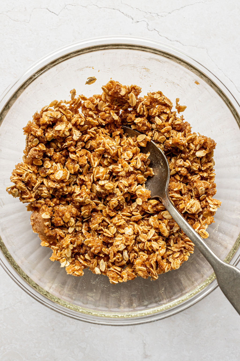 A glass bowl filled with an oat and brown sugar mixture, with a metal spoon resting inside, on a light-colored surface.