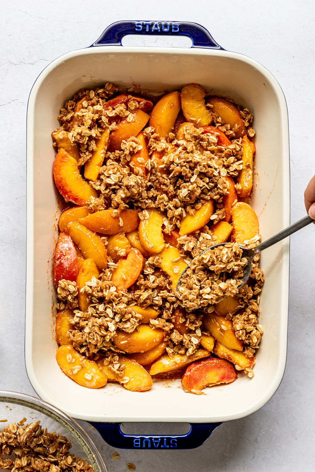A baking dish filled with sliced peaches topped with an oat crumble mixture, being spread evenly with a spoon.