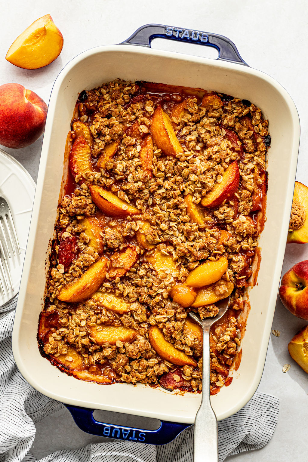 A baked peach crisp in a rectangular dish, with a serving spoon inside. Sliced peaches and an oat topping are visible. Fresh peaches and plates are nearby.