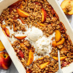 A baked peach crisp topped with two scoops of vanilla ice cream in a rectangular dish, surrounded by fresh peaches and a spoon.