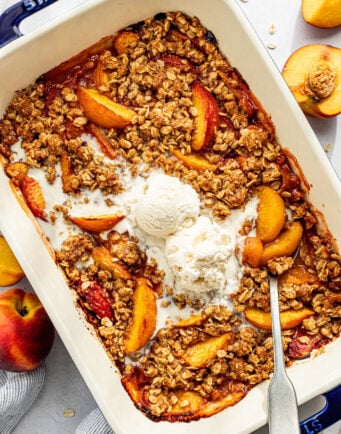 A baked peach crisp topped with two scoops of vanilla ice cream in a rectangular dish, surrounded by fresh peaches and a spoon.