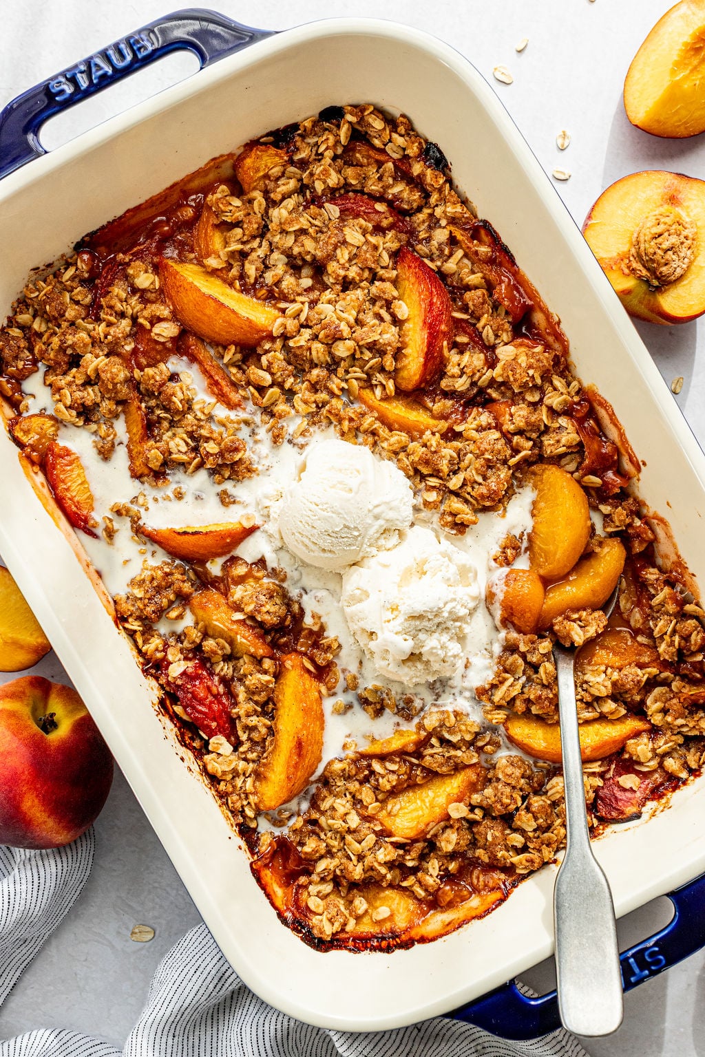 A baked peach crisp topped with two scoops of vanilla ice cream in a rectangular dish, surrounded by fresh peaches and a spoon.