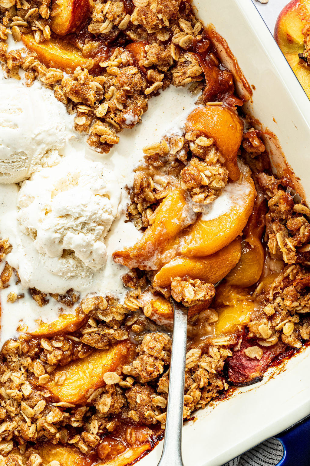 A close-up of peach crisp in a baking dish, topped with scoops of vanilla ice cream and a serving spoon beside baked peaches and oat crumble.