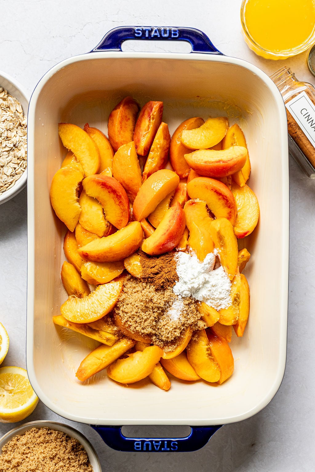 Sliced peaches, brown sugar, and flour are placed in a rectangular baking dish, surrounded by bowls of oats, brown sugar, lemon, juice, and a jar of cinnamon.