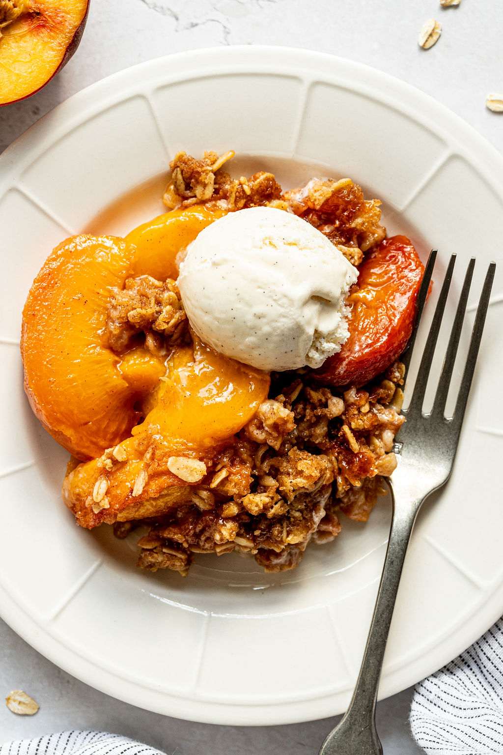 A serving of peach crisp topped with a scoop of vanilla ice cream on a white plate with a fork.