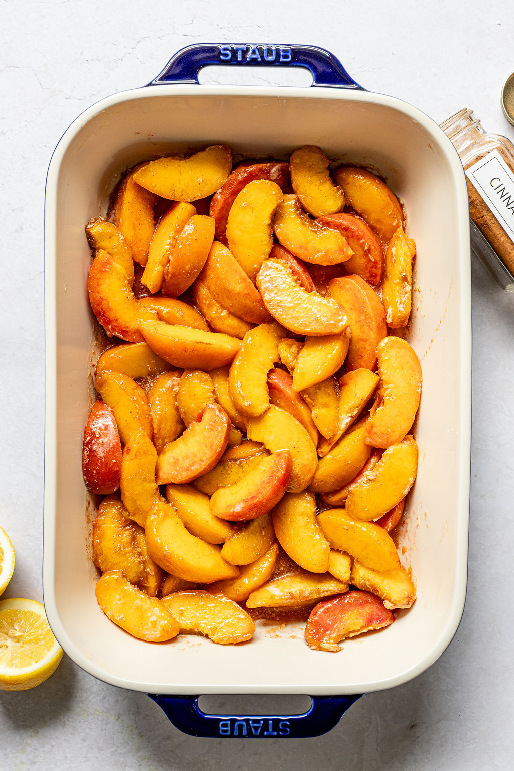 Sliced peaches coated with sugar and seasoning in a blue baking dish, with halved lemons and a jar of cinnamon nearby.