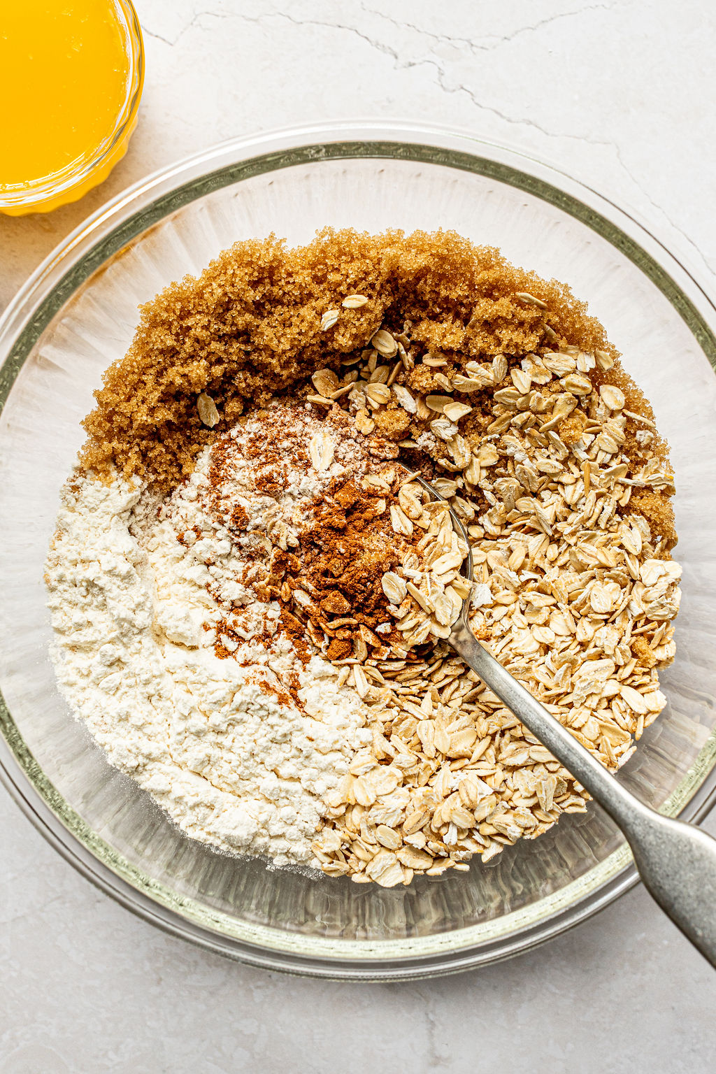 A glass bowl contains brown sugar, rolled oats, flour, and spices with a metal spoon; a bowl of melted butter is nearby on the counter.