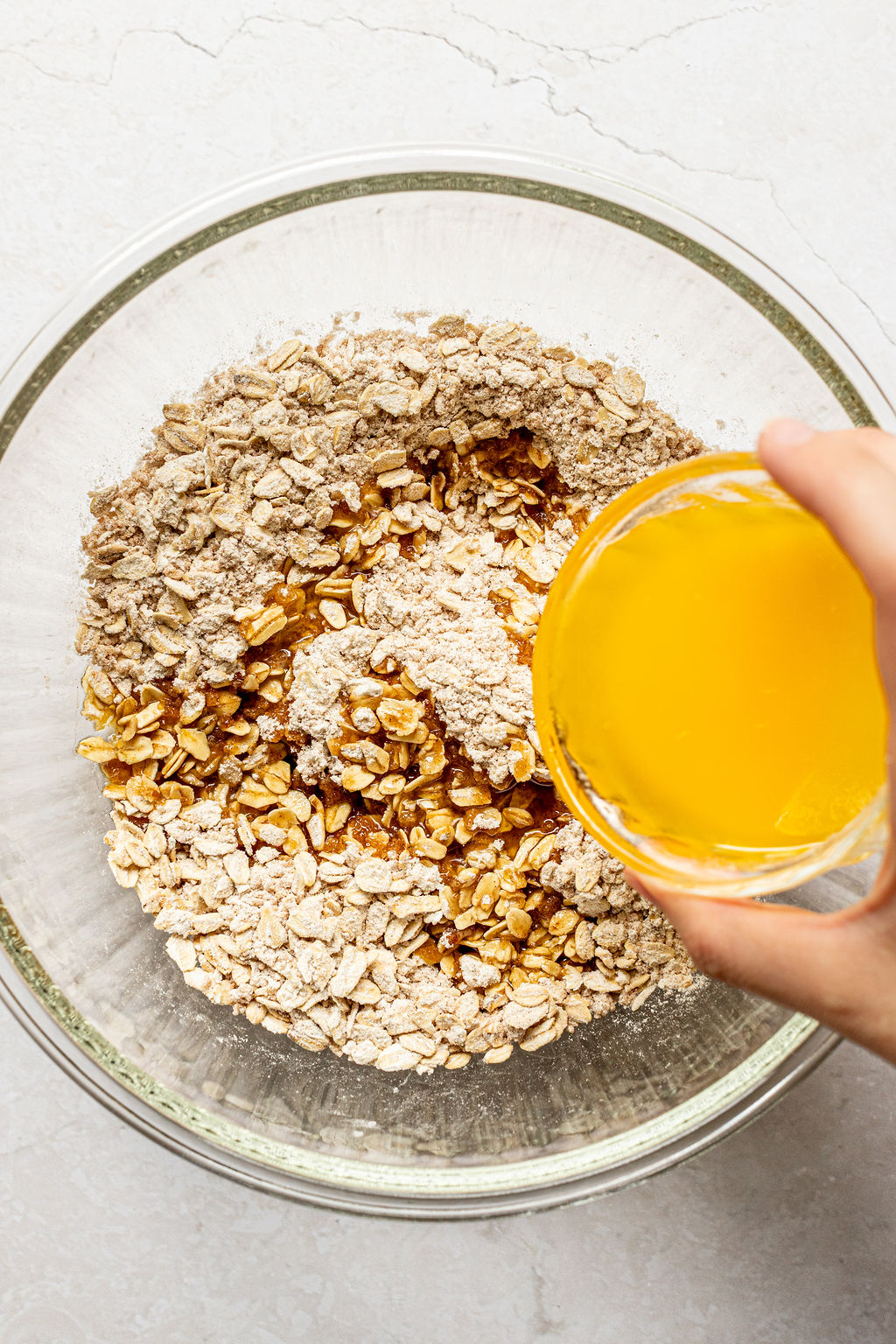 A hand pours melted butter from a small glass bowl into a larger bowl containing oats and flour on a light countertop.