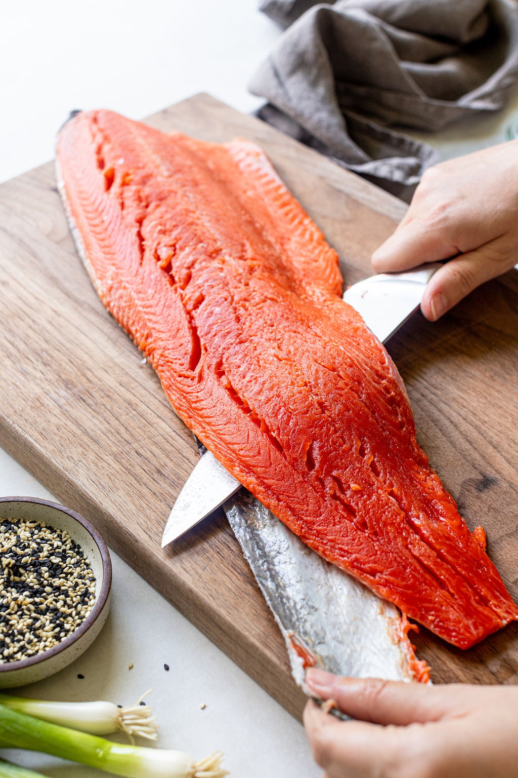 A person uses a knife to remove the skin from a large fillet of raw salmon on a wooden cutting board. A bowl of sesame seeds and green onions are nearby.