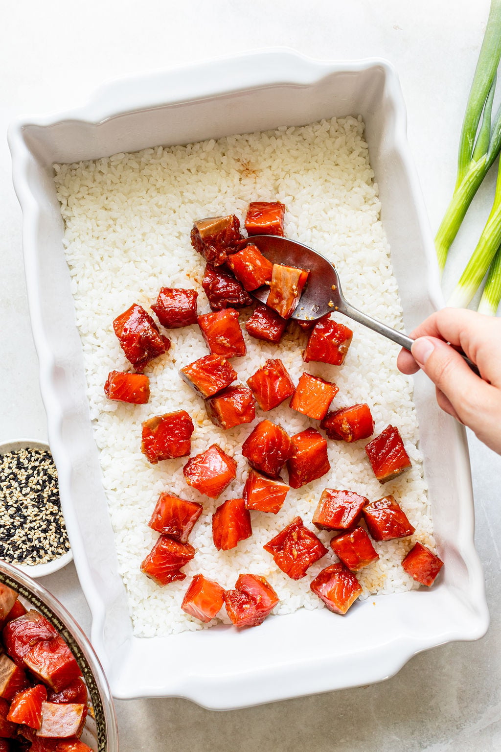 A hand spreads glazed salmon cubes over a layer of white rice in a rectangular baking dish; green onions and sesame seeds are nearby.