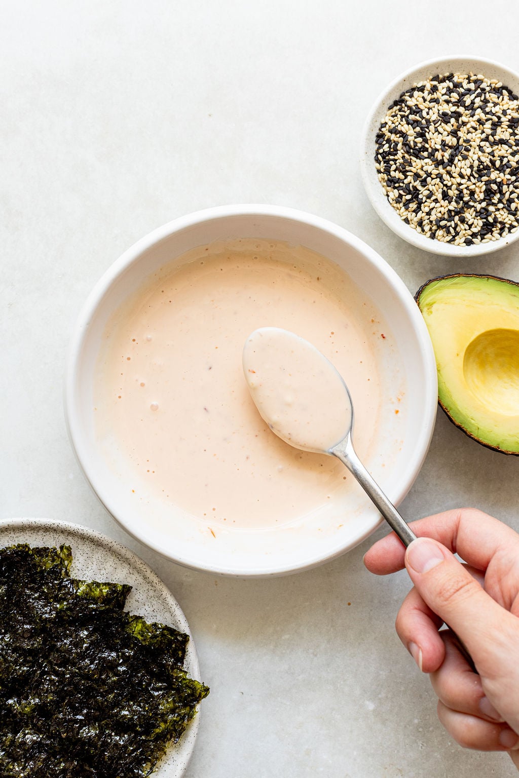 A hand holding a spoon over a bowl of creamy sauce, with nori sheets, half an avocado, and a bowl of sesame seeds nearby on a light surface.