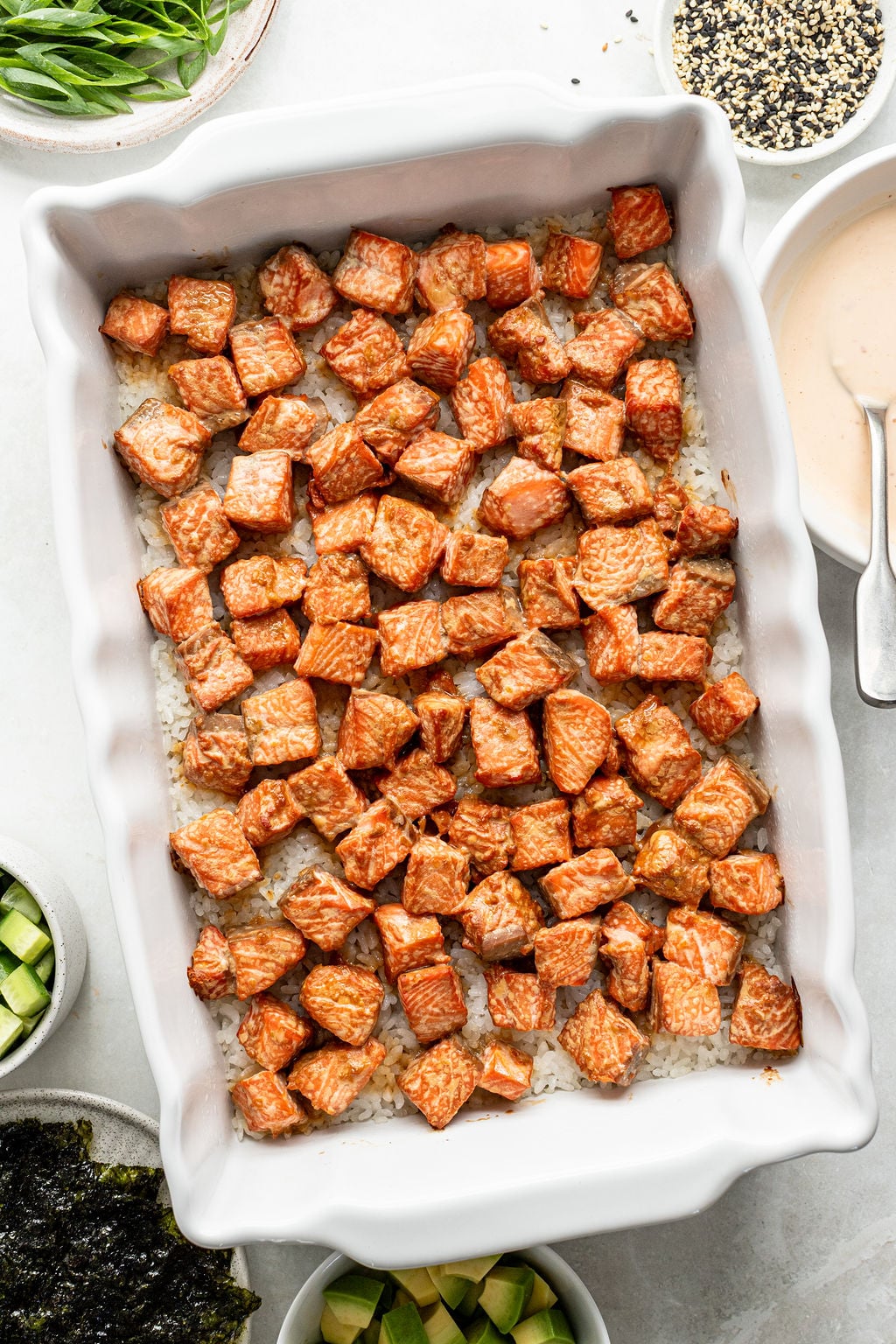 Cubed salmon coated in sauce is arranged on a layer of rice in a white baking dish, surrounded by bowls of sliced vegetables, sesame seeds, and sauce.