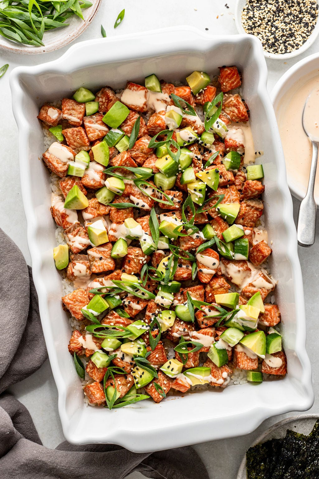 A white casserole dish filled with a deconstructed sushi bake, topped with diced salmon, avocado, green onions, sesame seeds, and drizzle of sauce.