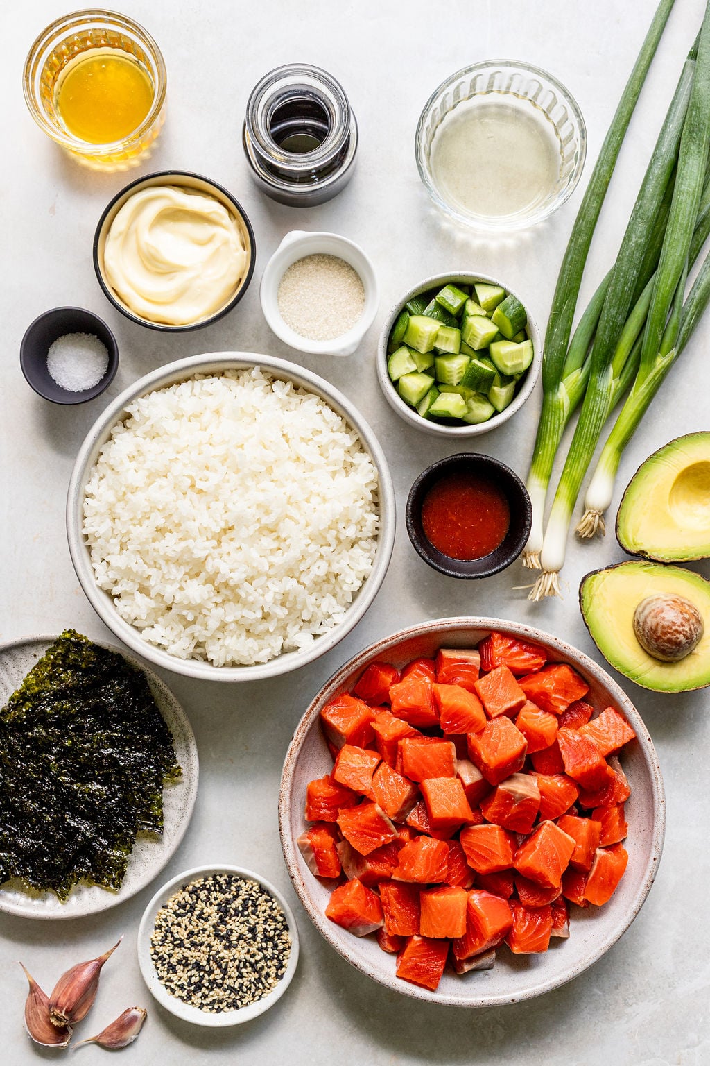 Various poke bowl ingredients are neatly arranged on a countertop, including diced raw salmon, rice, cucumber, avocado, scallions, nori, sauces, seasonings, and sesame seeds.