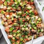 A casserole dish filled with a salmon poke bake topped with diced avocado, sliced green onions, sesame seeds, and a creamy sauce; a serving spoon is in the dish.