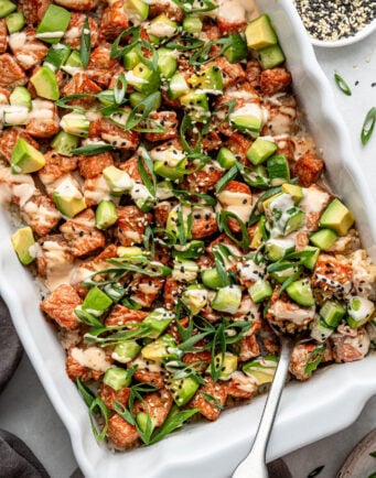 A casserole dish filled with a salmon poke bake topped with diced avocado, sliced green onions, sesame seeds, and a creamy sauce; a serving spoon is in the dish.