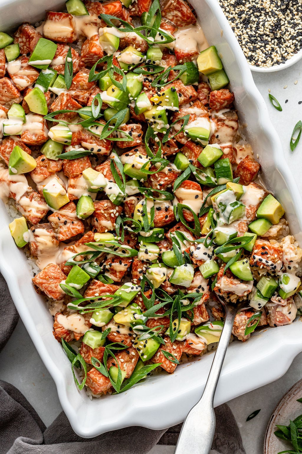 A casserole dish filled with a salmon poke bake topped with diced avocado, sliced green onions, sesame seeds, and a creamy sauce; a serving spoon is in the dish.