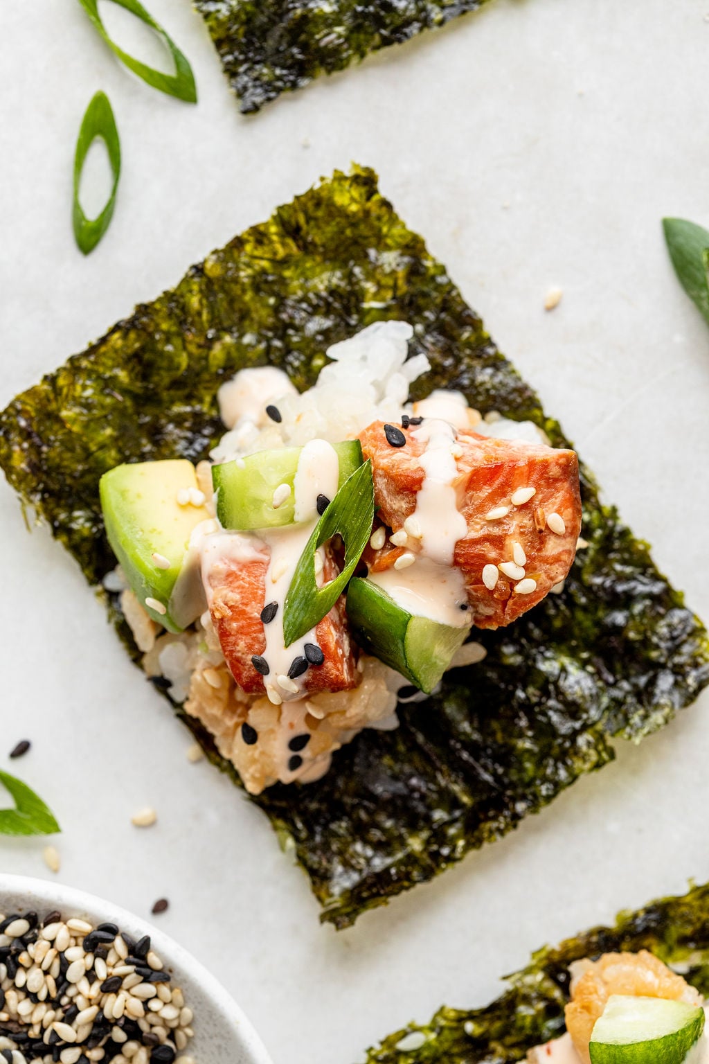 A piece of seaweed topped with sushi rice, avocado, salmon, sliced green onion, sesame seeds, and a drizzle of creamy sauce.