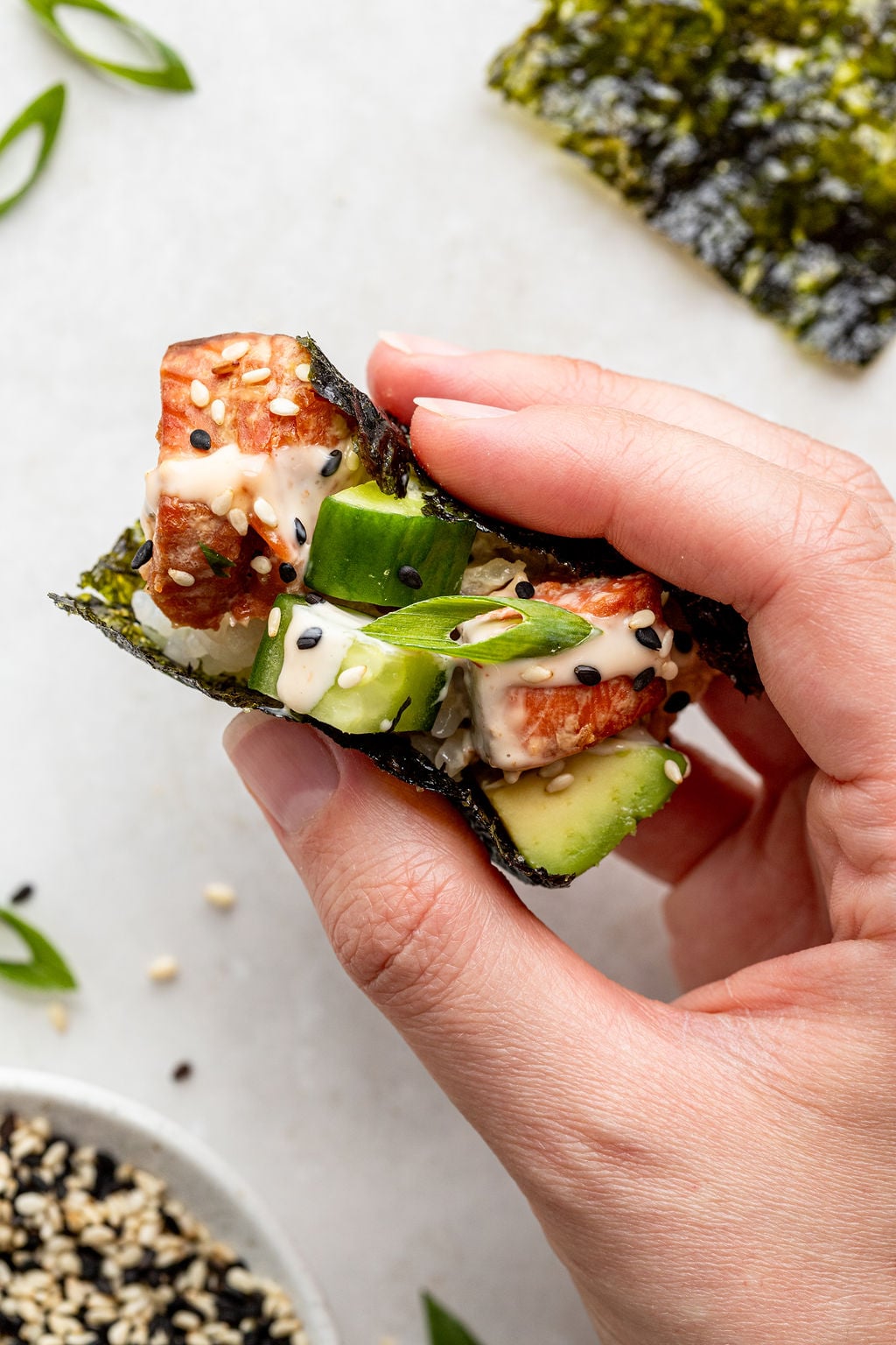 A hand holds a piece of seaweed wrapped around cubes of fish, cucumber slices, green onion, and a creamy sauce, topped with black and white sesame seeds.