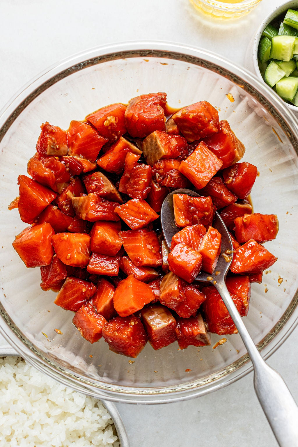 A glass bowl filled with marinated raw salmon cubes, with a spoon resting inside. Nearby are bowls of white rice and chopped cucumber.