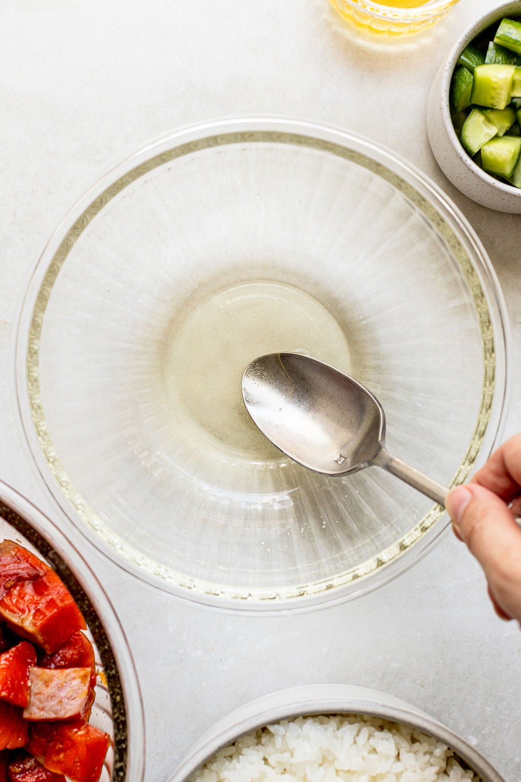 A hand holds a spoon over a glass mixing bowl with liquid at the bottom, surrounded by chopped salmon, cucumbers, and a bowl of rice.