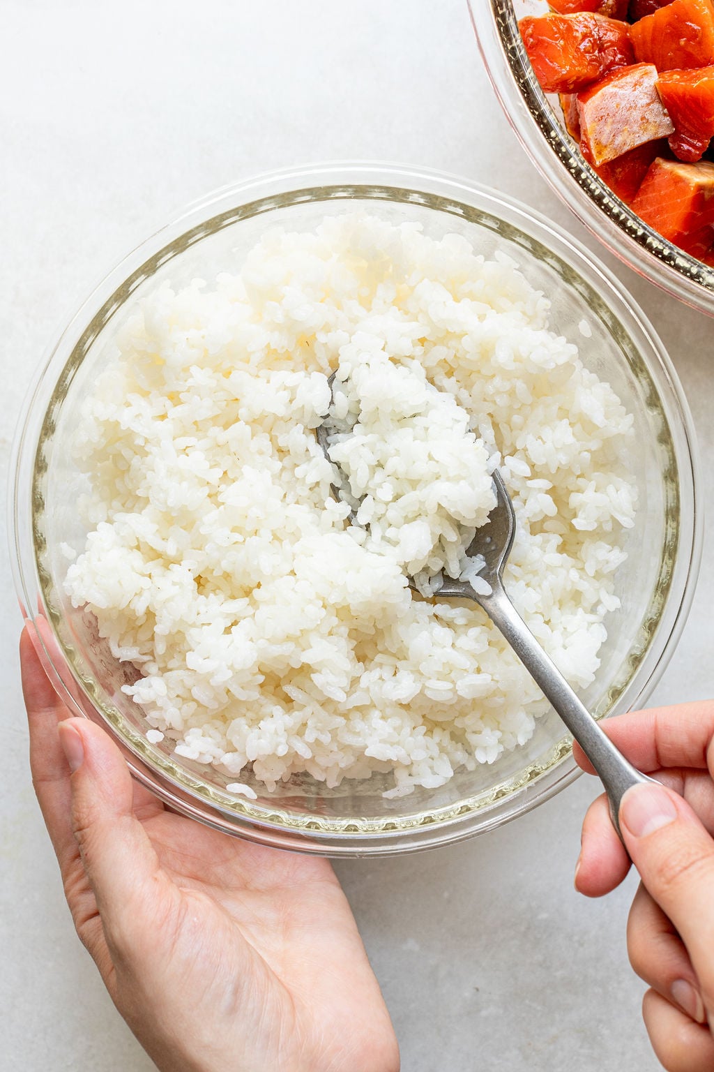 A hand holds a bowl of cooked white rice while another hand stirs it with a spoon; a bowl of seasoned fish is partially visible in the corner.