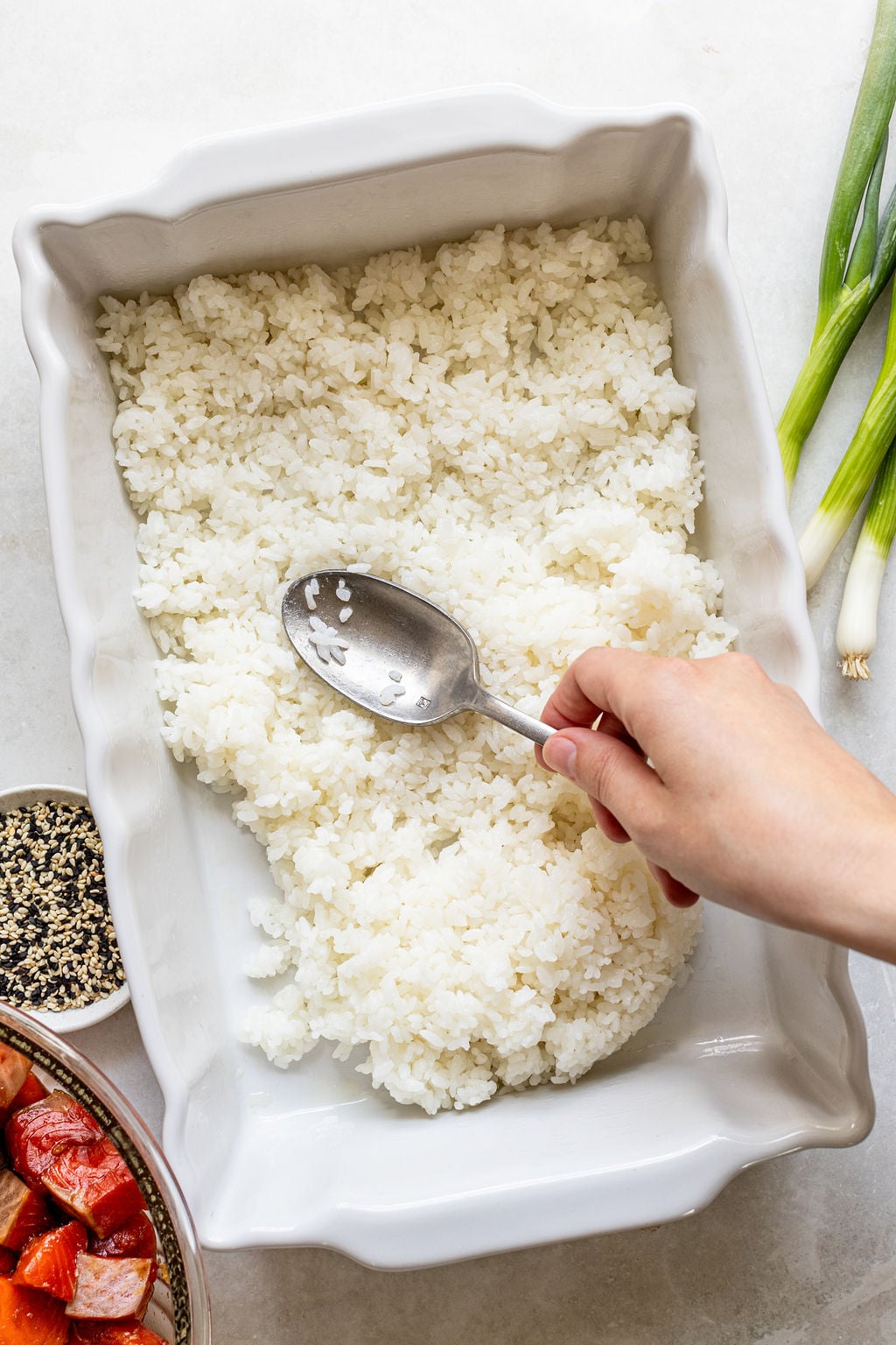 A hand spreads cooked rice evenly in a white baking dish with a spoon. Green onions, sesame seeds, and cubed salmon are visible nearby.